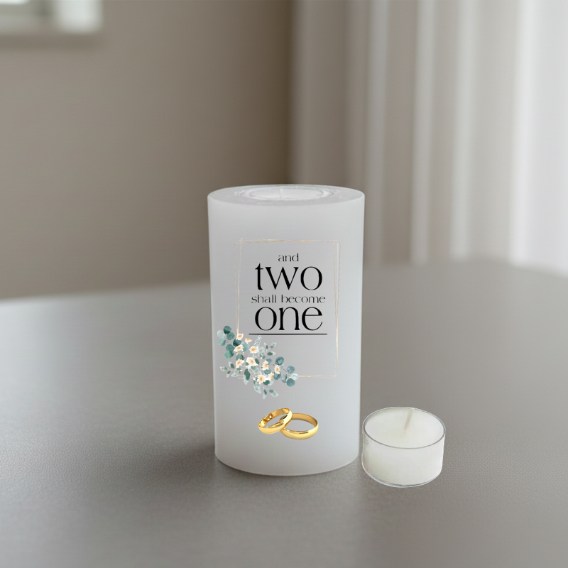 A white candle on a grey tabletop, printed with the phrase 'and two shall become one' in black serif text, a small cluster of pale blue and white flowers, and two interlocking gold rings near the base.