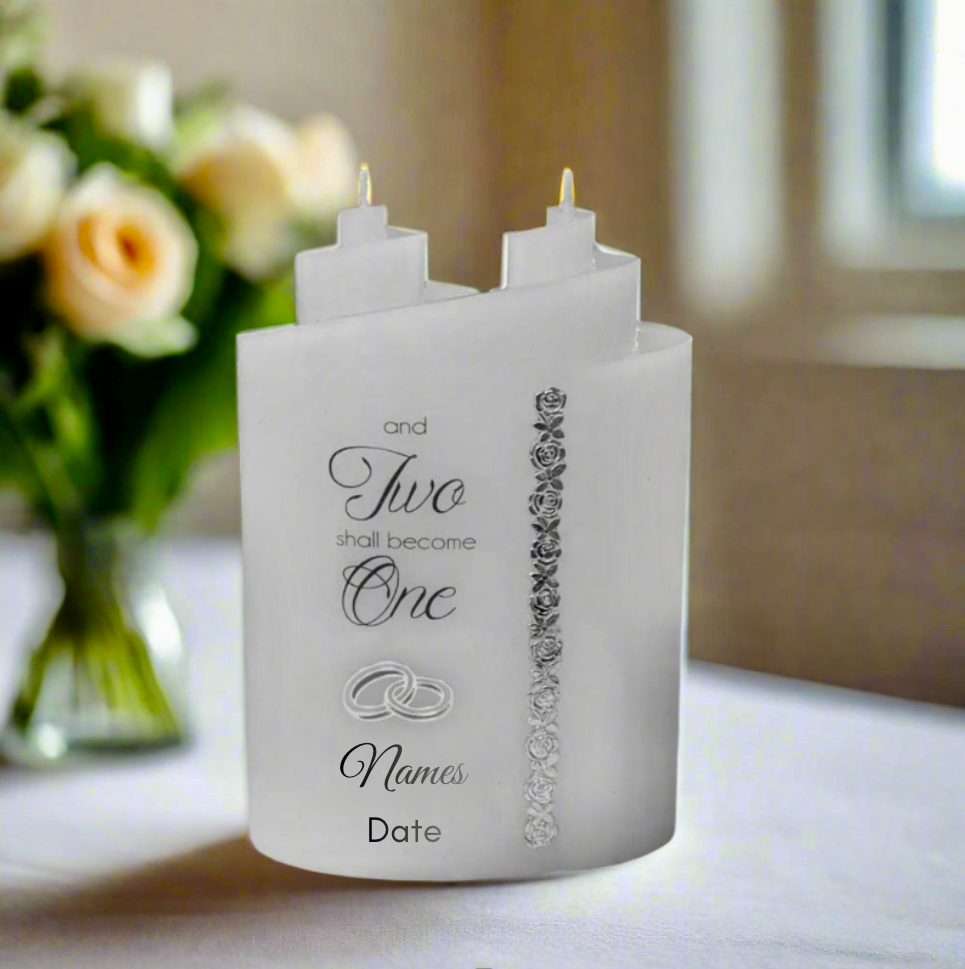 White candle with 2 wicks and engraved text and decorative elements on a table with flowers in the background.