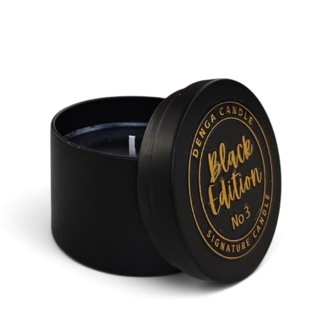 A black round tin candle with its lid leaning against it on a white background. The lid features gold foil text reading 'DENGA CANDLE Black Edition No3