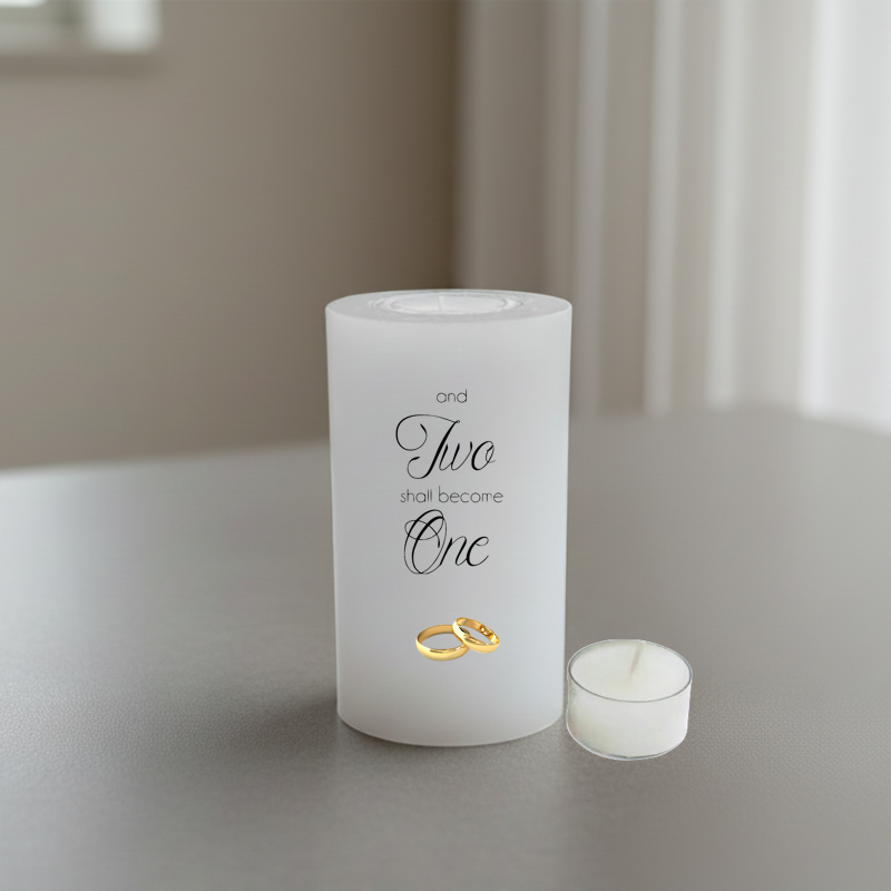 A white candle with black script text reading 'and Two shall become One' and two small gold rings, placed on a grey surface.