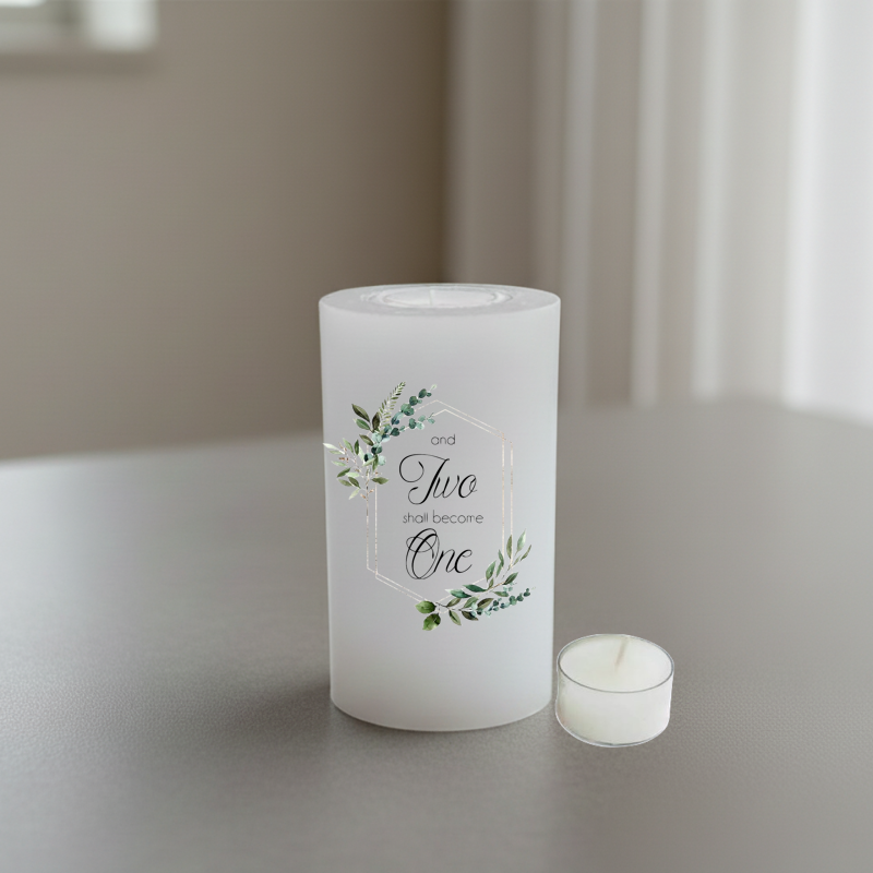 A white cylindrical candle with a printed floral wreath and the words 'and Two shall become One' stands on a tabletop next to a small white tea light candle.