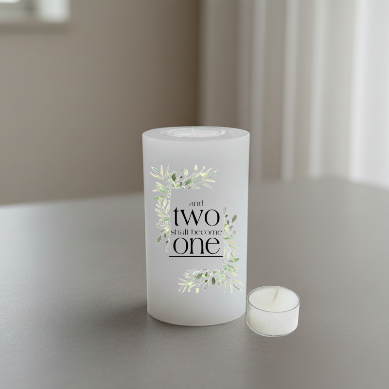A white candle with a printed design that reads 'and two shall become one' in black serif text, surrounded by delicate green and white floral illustrations.