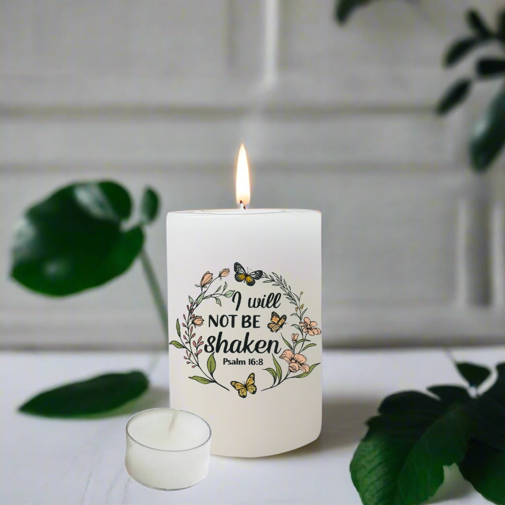 A white pillar candle with a lit flame and a small matching tealight candle on a light surface, printed with the phrase 'I will NOT BE shaken Psalm 56:8' inside a floral wreath design with butterflies.