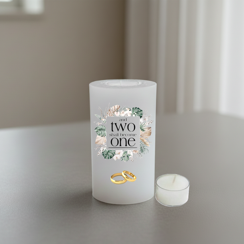 white candle with a floral graphic and the text 'and two shall become one' on the front, shown standing on a grey surface with a softly blurred indoor background.