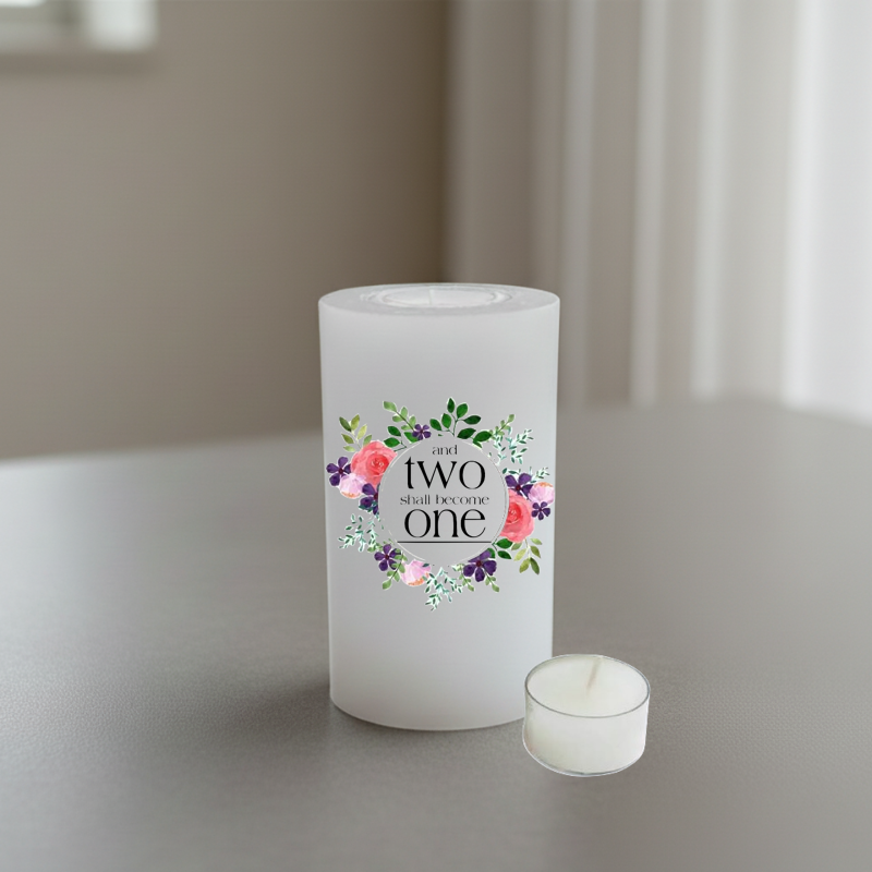 A white cylindrical candle standing on a tabletop, printed in black script and surrounded by a floral wreath in pink, purple, green, and white.