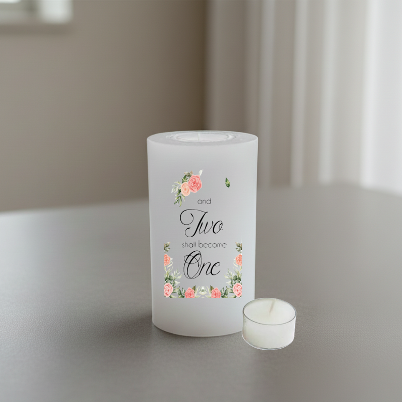 A white wedding candle with a floral design and text printed on the front, set on a grey tabletop.
