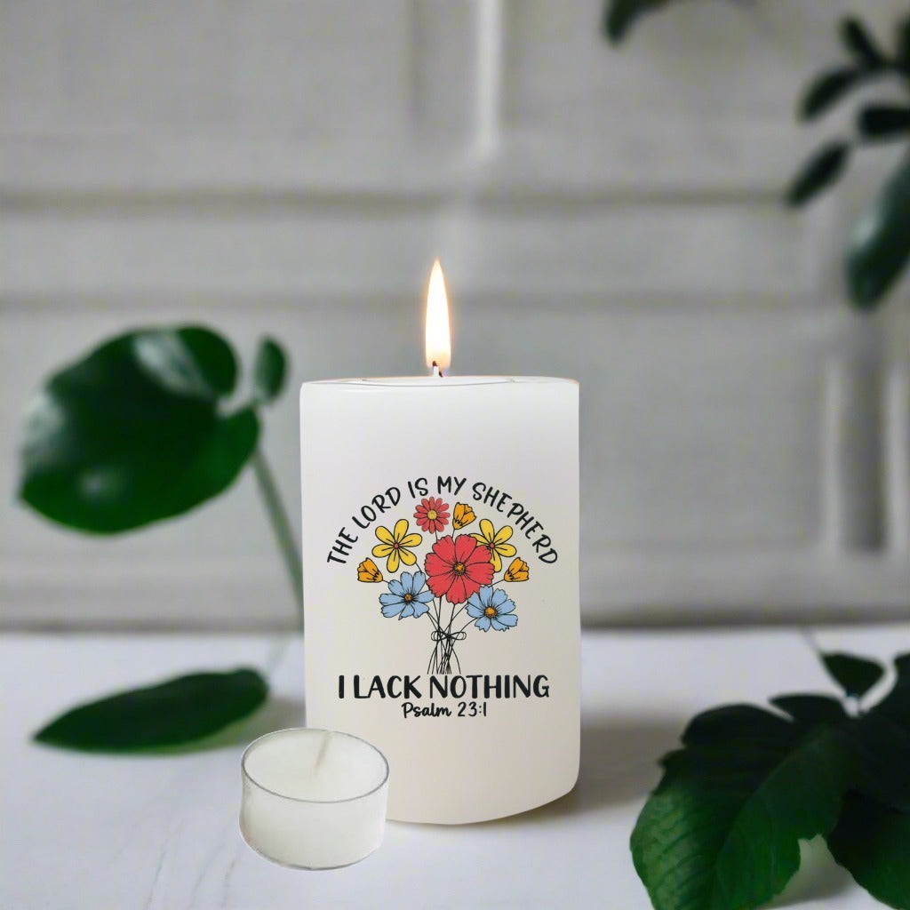 A lit white candle with a printed floral design and the text 'THE LORD IS MY SHEPHERD I LACK NOTHING Psalm 23:1' on the front, styled on a white surface with green leaves in the background.