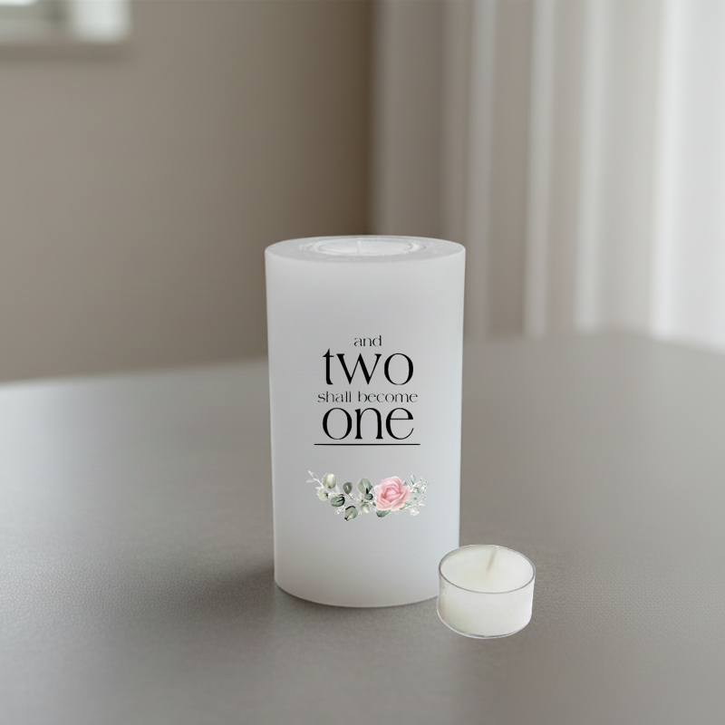 A white cylindrical candle with the printed phrase 'and two shall become one' in black serif text, plus a small pink and white floral illustration near the bottom.