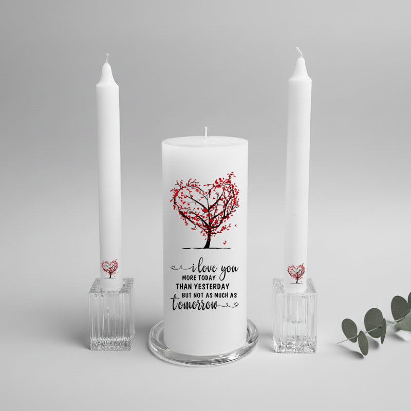 White candle with red heart design and text, flanked by two tall white candles on a light gray background.