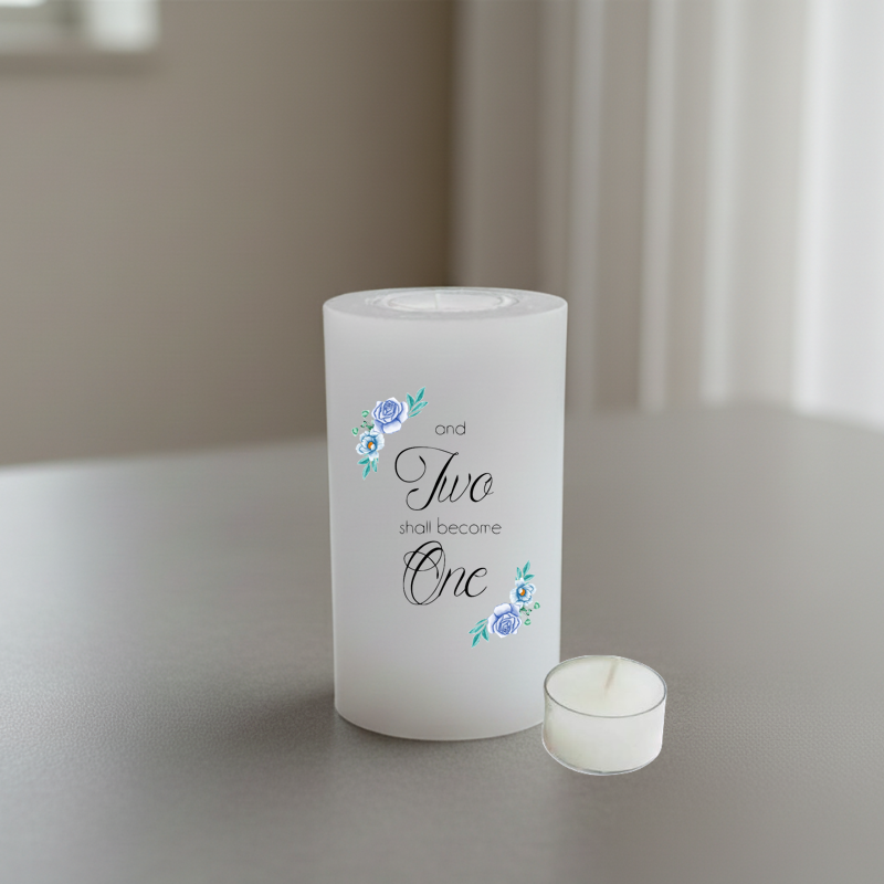 A white candle with printed text reading 'and Two shall become One' in black script and serif fonts, accented with small blue and green floral illustrations.