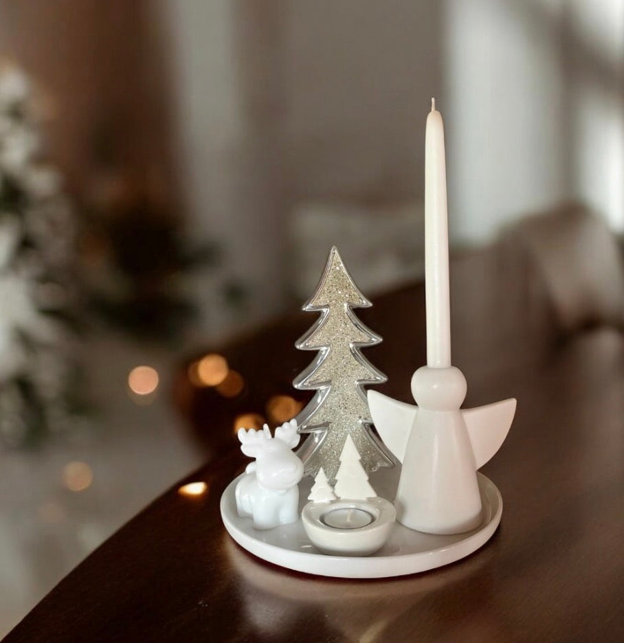 A white ceramic Christmas candle holder set on a dark wooden table, featuring a white angel candle holder with a tall white taper candle, a silver glitter Christmas tree, a small white reindeer figurine, and a tea light holder