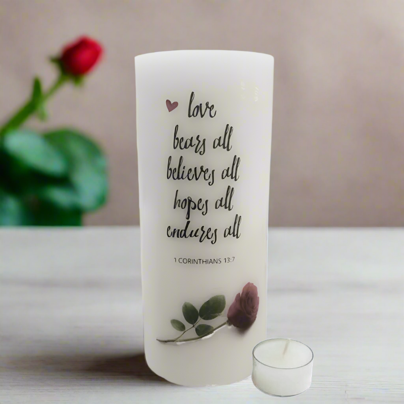 A white pillar candle with a Bible verse from 1 Corinthians 13:13 printed on it, decorated with a small rose and green leaves.
