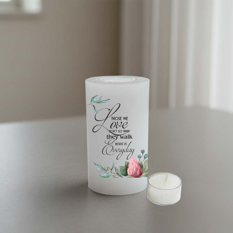 A white candle printed black script text and pink floral illustrations, standing on a grey tabletop.