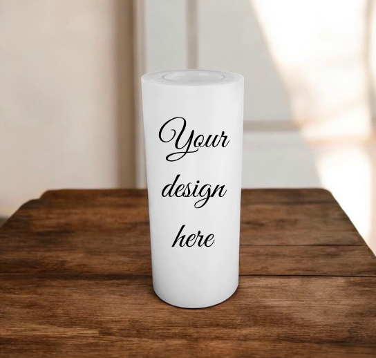A white candle with the words 'Your design here' printed in black script, standing on a wooden tabletop against a softly blurred indoor background.