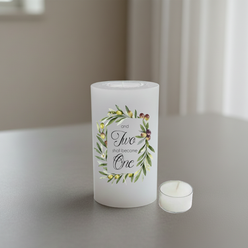 A white candle with a botanical wreath design and the printed phrase “and Two shall become One,” shown on a tabletop in a softly blurred indoor setting.