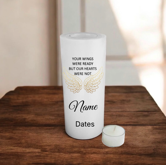 A white memorial candle with printed text and angel wings design sits on a wooden table