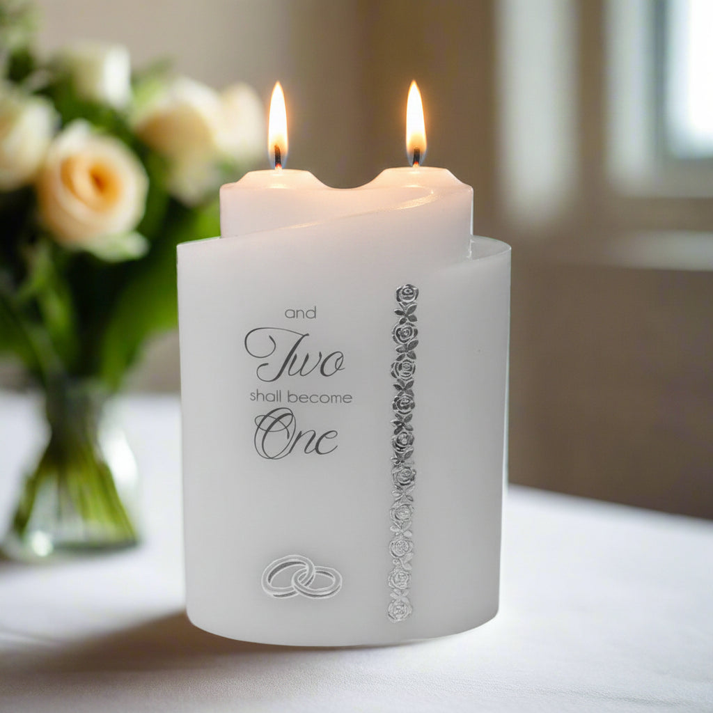 Two white candles with decorative text and design on a table with flowers in the background