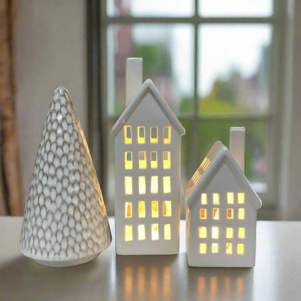 two porcelain tealight houses with lights inside and one ceramic tree on a window sill