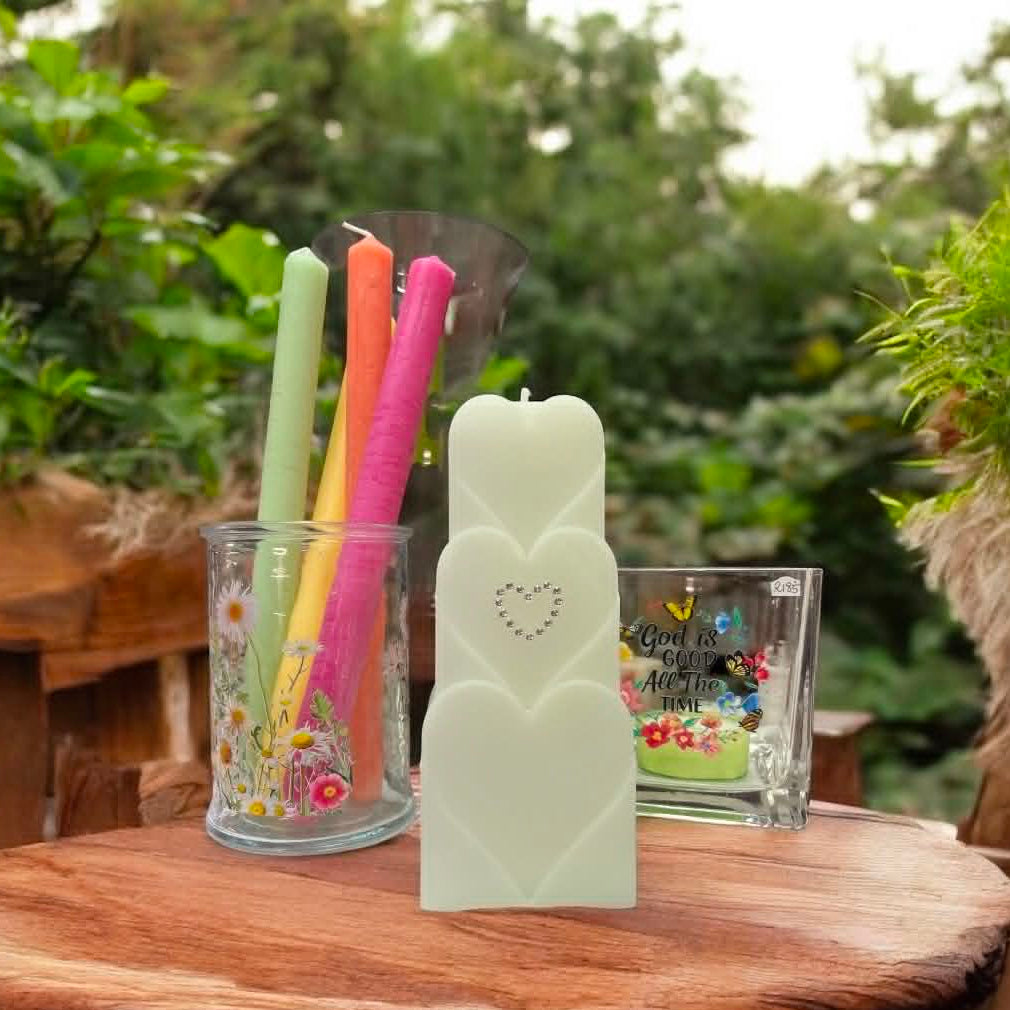 A heart-shaped white candle displayed on a rustic wooden table outdoors, surrounded by green foliage. 