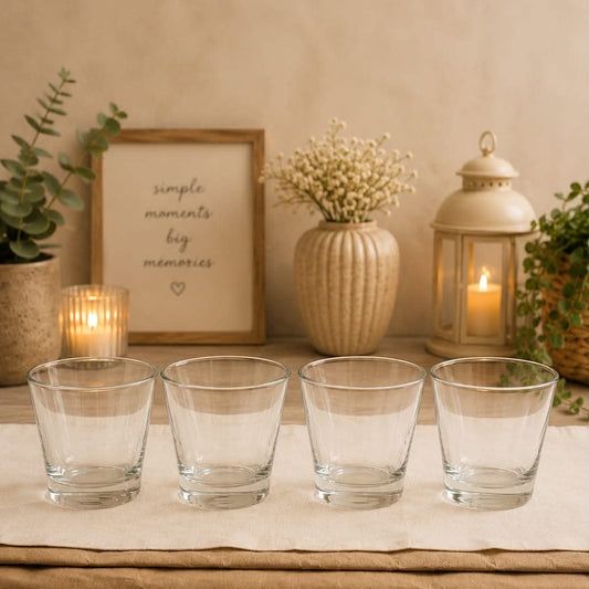 Four clear glass tumblers arranged in a row on a beige runner, styled on a wooden table with candles, a framed sign, and decorative plants in the background.