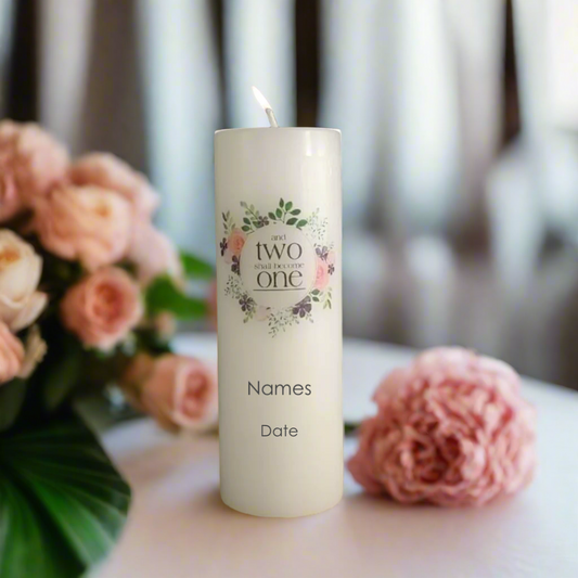 A personalised white pillar candle with a lit wick stands on a table, decorated with a floral wreath and the text, along with placeholder names and date.