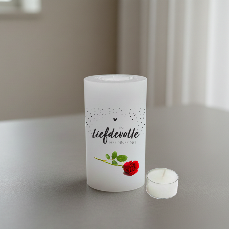 A white candle with black Afrikaans text reading and a red rose illustration, on a grey surface