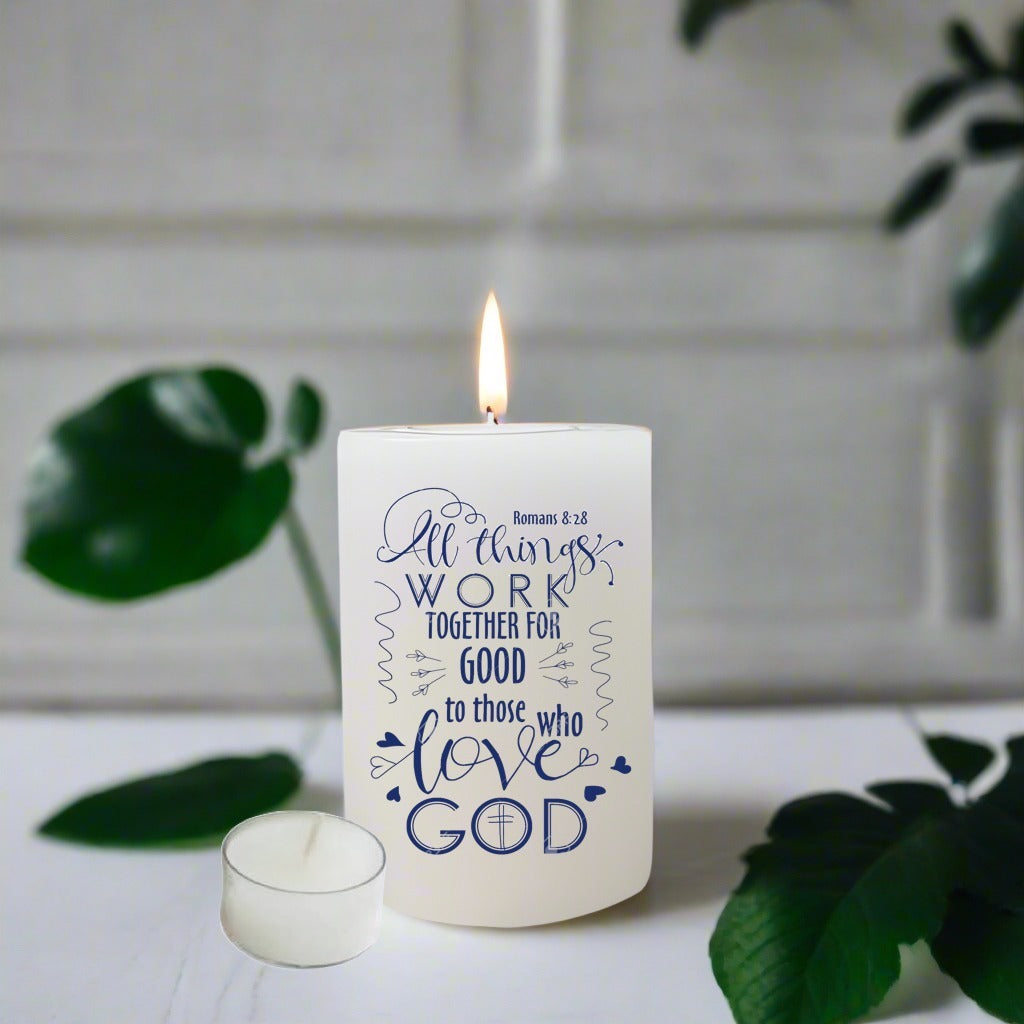 Candle with text 'All things work together for good to those who love God' on a white surface with green leaves.