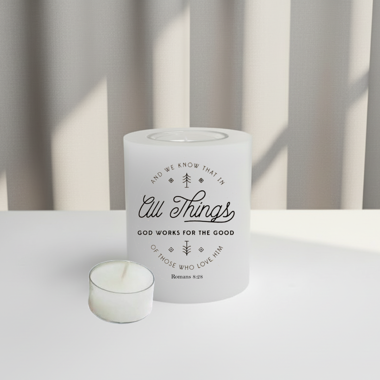 White candle with text Romans 8:28 on a white surface and a tealight on the side.