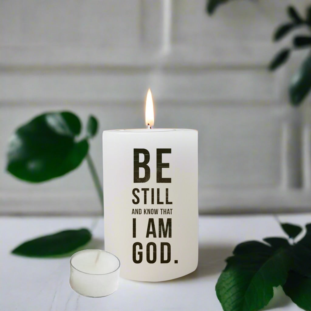 Candle with 'Be Still and Know That I Am God' text on a white surface with green leaves.