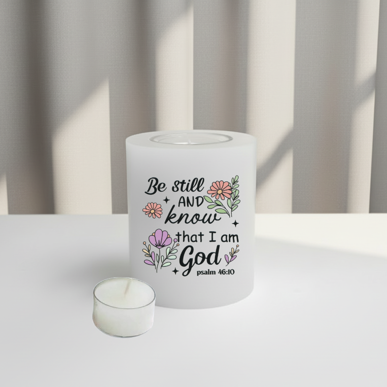 A white candle with a printed Bible verse and floral illustrations, sitting on a white surface with a soft-focus background of vertical blinds.