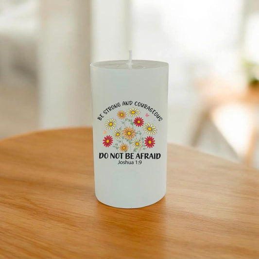 A white pillar candle with a printed design featuring the text 'BE STRONG AND COURAGEOUS DO NOT BE AFRAID Joshua 1:9' and a cluster of daisy-like flowers in yellow, red, and white, sitting on a wooden table.