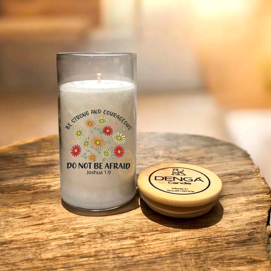 A glass jar candle with label reads 'BE STRONG AND COURAGEOUS DO NOT BE AFRAID Joshua 1:9' with small red and yellow flowers.