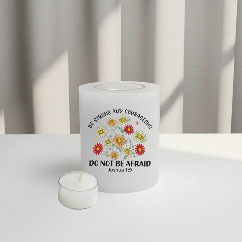 A  white candle with a printed design featuring colorful daisies and the text 'BE STRONG AND COURAGEOUS DO NOT BE AFRAID Joshua 1:9' on a white surface with a soft-focus background.