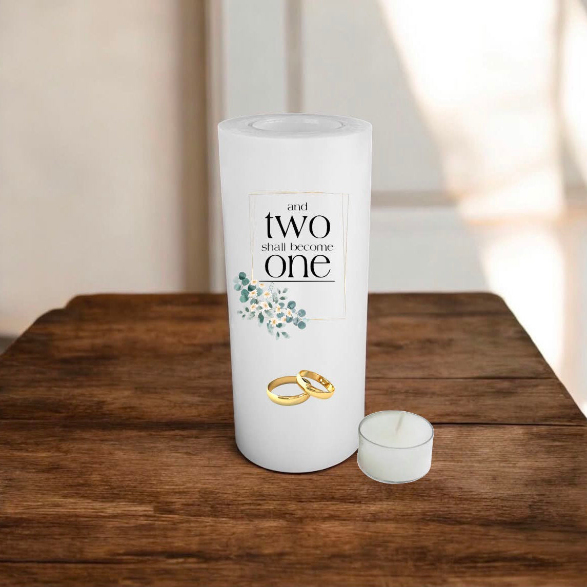 A white candle with a custom printed label reading 'and two shall become one' with floral accents and two gold rings, displayed on a rustic wooden table.