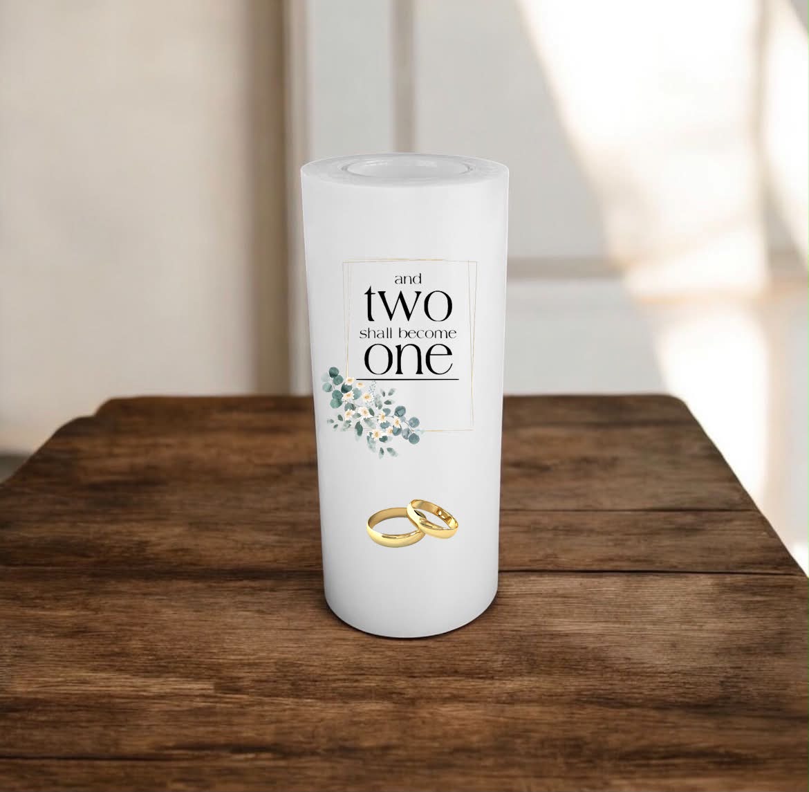 A white candle with a custom printed label reading 'and two shall become one' with floral accents and two gold rings, displayed on a rustic wooden table.