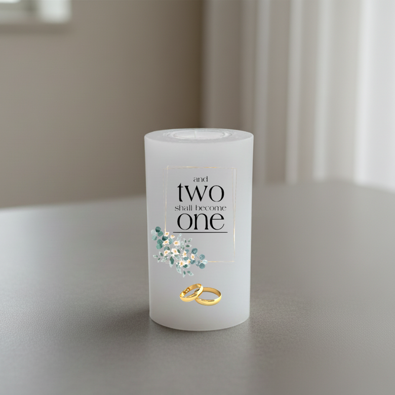 A white candle on a grey tabletop, printed with the phrase 'and two shall become one' in black serif text, a small cluster of pale blue and white flowers, and two interlocking gold rings near the base.