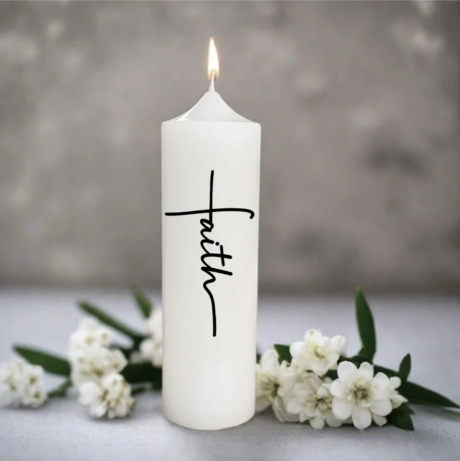 A white pillar candle with the word 'faith' written in black cursive script, shown lit with small white flowers and green leaves at its base.