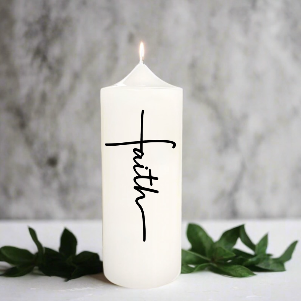 A white pillar candle with the word 'faith' written in black cursive script, lit with a small flame, set on a white surface with green leaves.