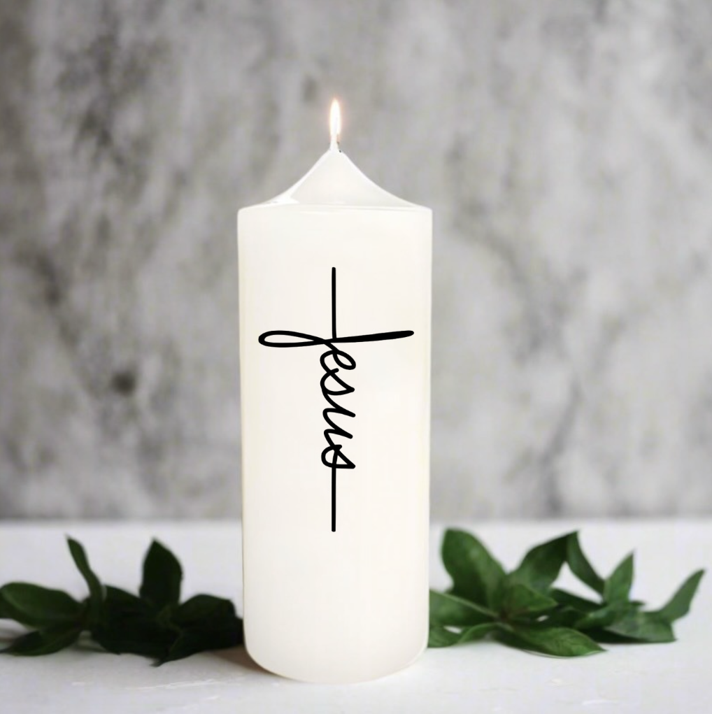 A white pillar candle with a lit flame, featuring the word 'Jesus' in black cursive script on the front, styled with green leaves