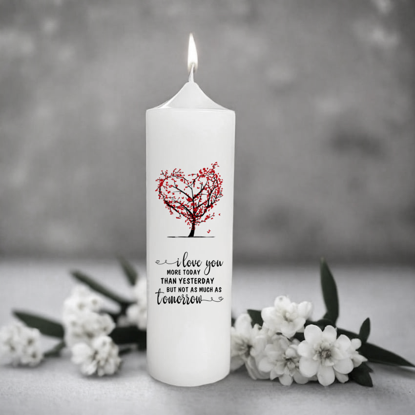 A white candle featuring a red heart-shaped tree design and the printed message 'I love you MORE TODAY THAN YESTERDAY BUT NOT AS MUCH AS tomorrow'.