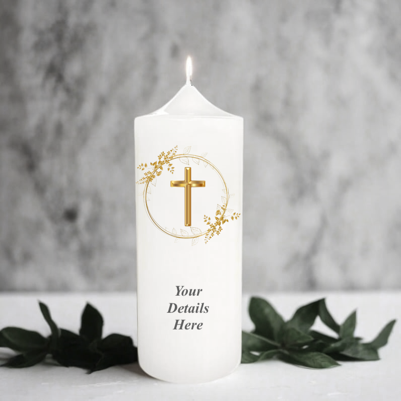 A  white pillar candle with a lit flame, featuring a gold cross inside a circular floral design and the text.