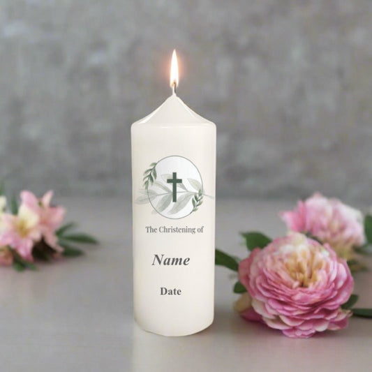 White christening candle with a cross design and text, surrounded by pink flowers on a grey background