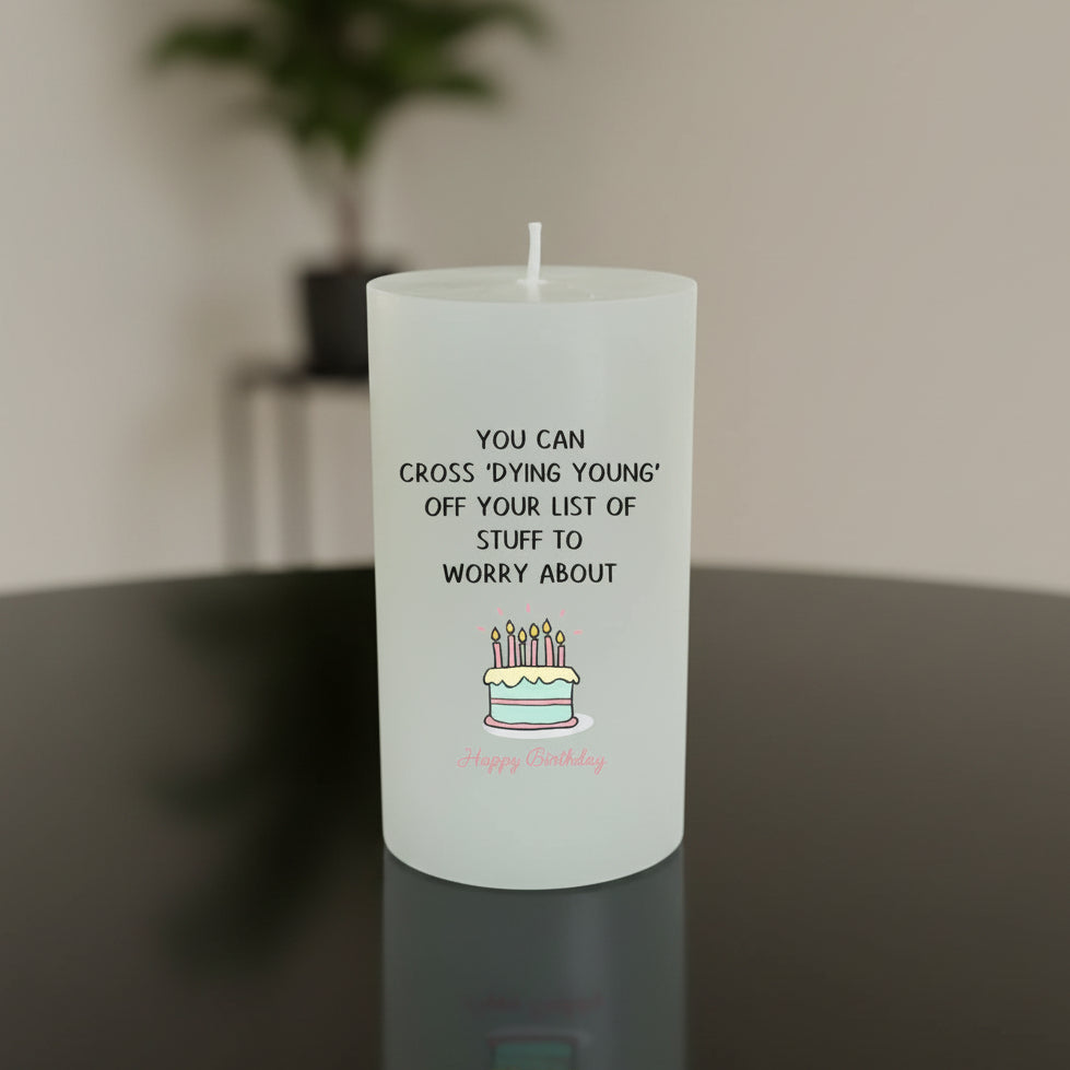 A white candle with black text reading 'YOU CAN CROSS DYING YOUNG! OFF YOUR LIST OF STUFF TO WORRY ABOUT' and a small birthday cake illustration, sitting on a wooden table with a blurred indoor background.