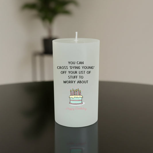 A white candle with black text reading 'YOU CAN CROSS DYING YOUNG! OFF YOUR LIST OF STUFF TO WORRY ABOUT' and a small birthday cake illustration, sitting on a wooden table with a blurred indoor background.