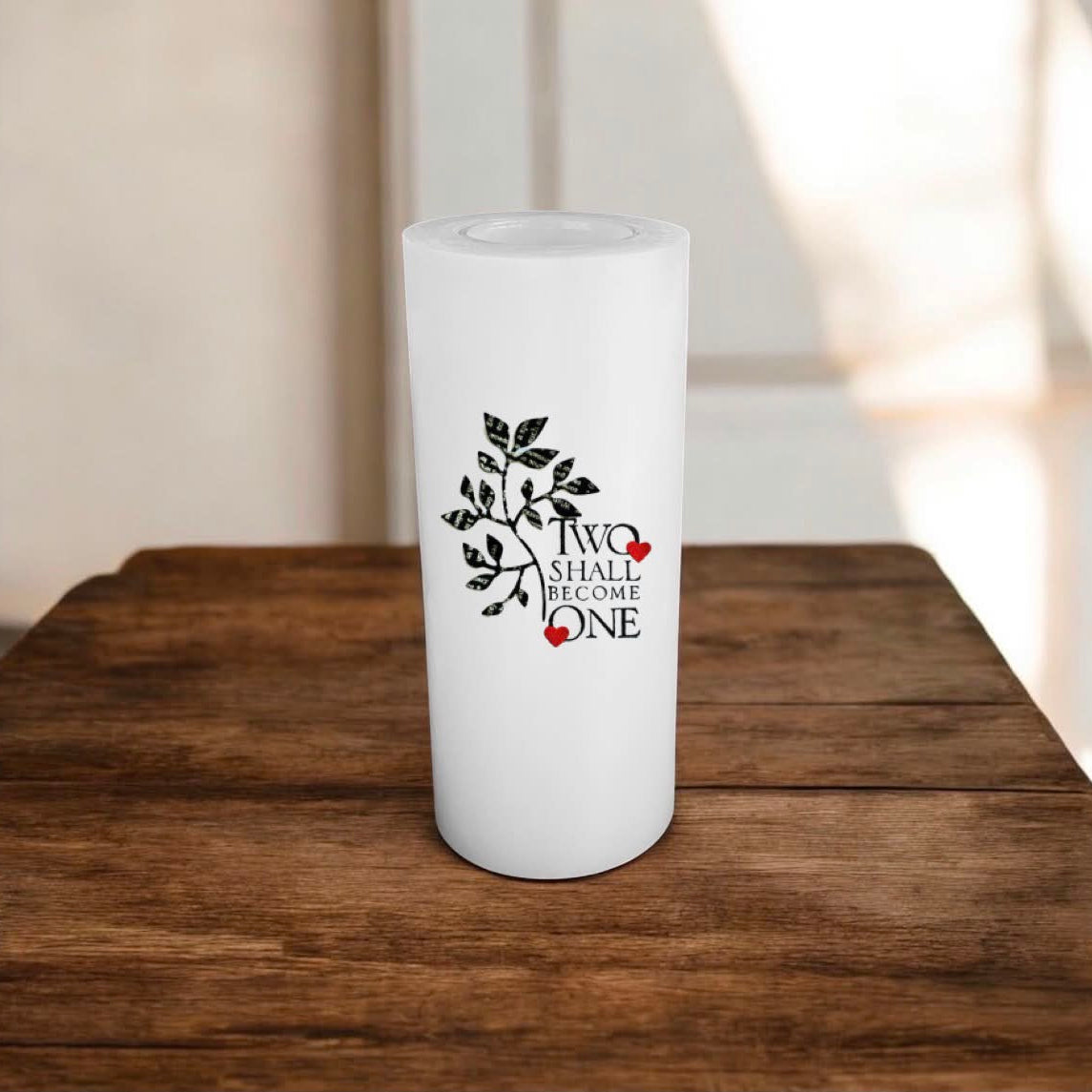 A white candle with a black leaf graphic and the words 'TWO SHALL BECOME ONE' with small red heart accents, displayed on a rustic wooden table.