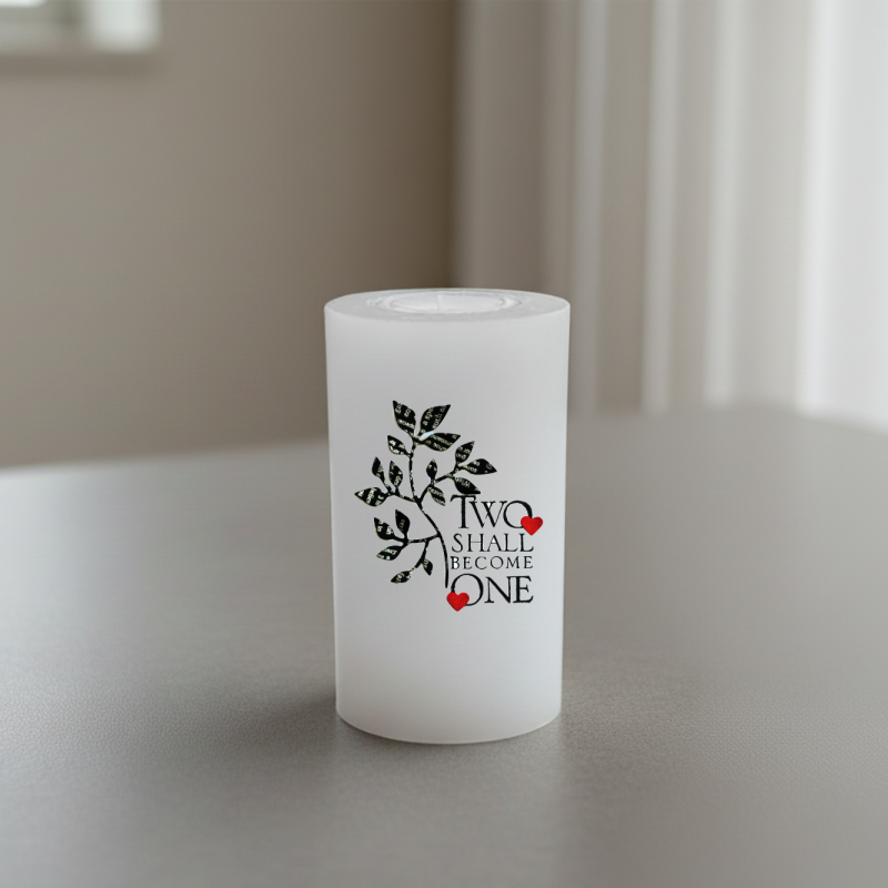 A white candle sits on a grey tabletop. The front features a black leafy branch graphic and the words 'TWO SHALL BECOME ONE' with small red hearts.