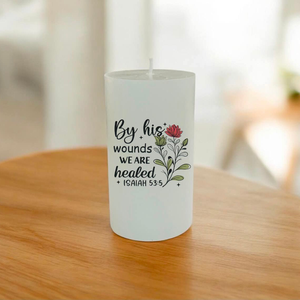 A white pillar candle with a printed Bible verse and floral design, sitting on a wooden table in a softly blurred indoor setting.