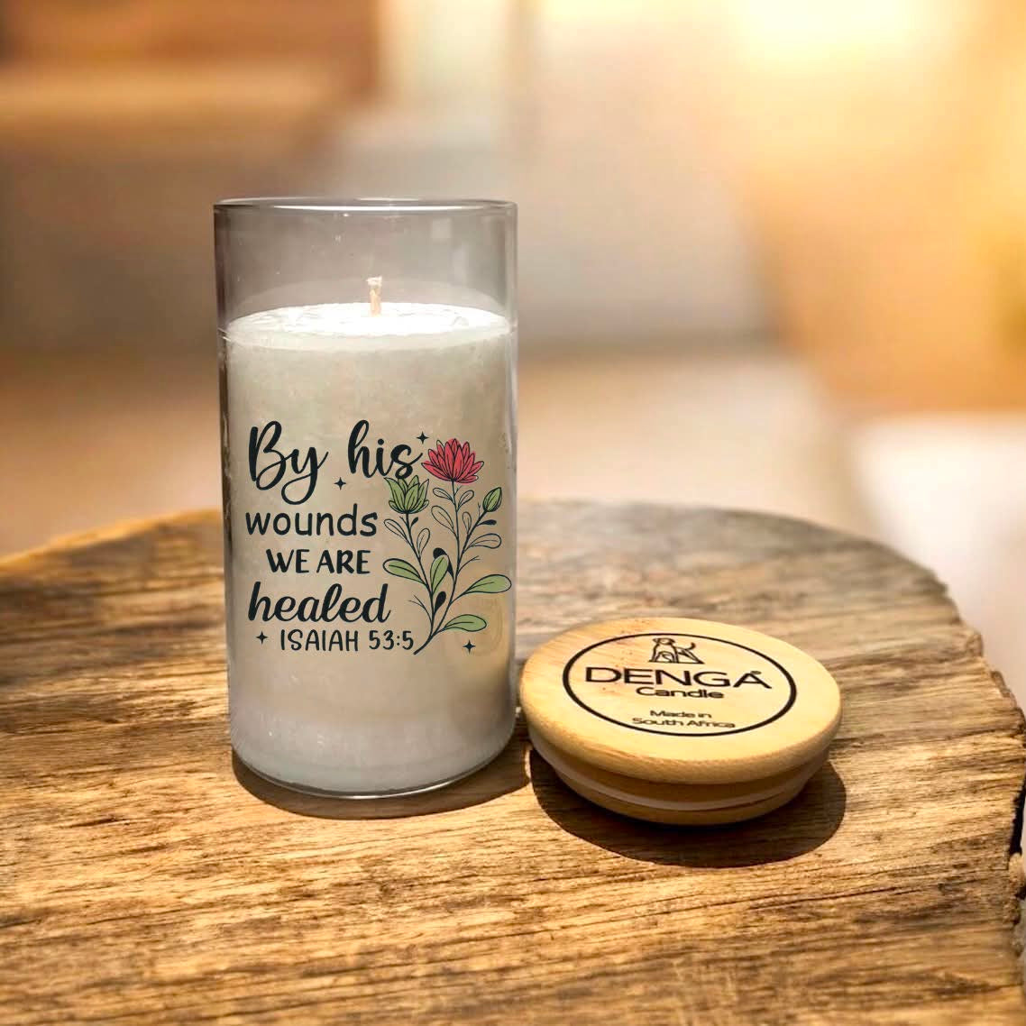 A glass jar candle with a printed quote reading 'By his wounds we are healed (Isaiah 53:5)' next to a round wooden lid .