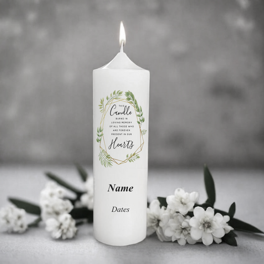 White memorial candle with engraved text and floral design on a gray background with white flowers.