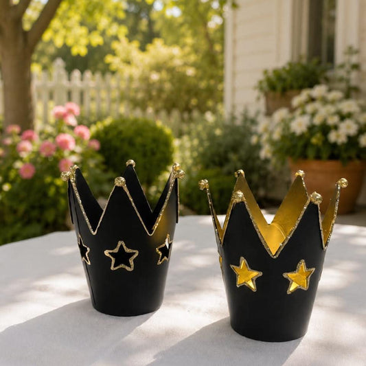 Two black crown-shaped candle holders with gold trim and star cutouts, displayed on a white tablecloth in an outdoor garden setting.
